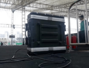 A large black equipment case with cables attached sits on a stage under a tent. Metal railings and a person in the background are visible. {{brizy_dc_image_alt imageSrc=