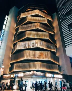A modern, multi-story building with illuminated, layered wooden slats on the facade at night; people walk by on the street below. {{brizy_dc_image_alt imageSrc=