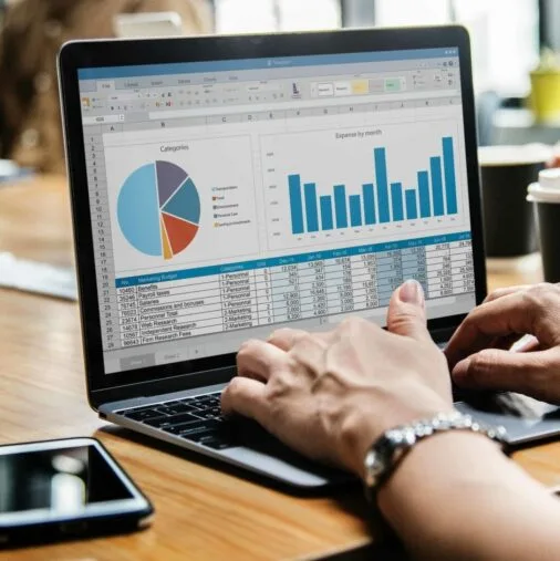 A person works on a laptop displaying spreadsheets with a pie chart and bar graph, while others sit around a table with papers, coffee cups, and a smartphone. {{brizy_dc_image_alt imageSrc=