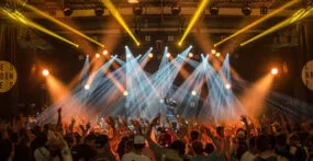 A large crowd with raised hands faces a stage with bright spotlights and a performer, in an indoor concert or music festival setting. {{brizy_dc_image_alt imageSrc=