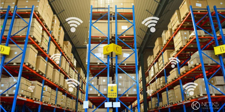 Large warehouse with tall shelves stacked with cardboard boxes; several Wi-Fi signal icons are overlaid, suggesting wireless connectivity throughout the space.